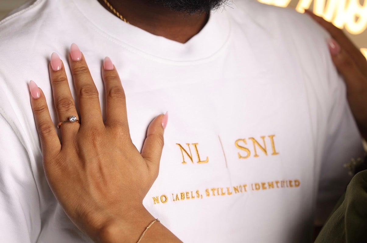 A close-up of a person wearing a white T-shirt with gold text reading 'NL SNI' and 'NO LABELS, STILL NOT IDENTIFIED'. A hand with pink manicured nails rests on the chest, and part of a ring is visible.


